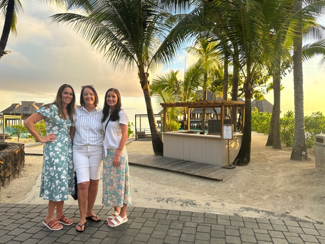 Three women standing in front of ocean and palm trees