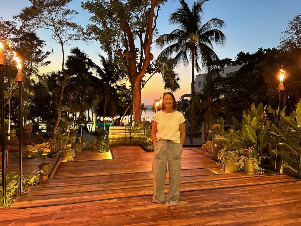 Woman standing on deck at sunset with palm trees and ocean in background.